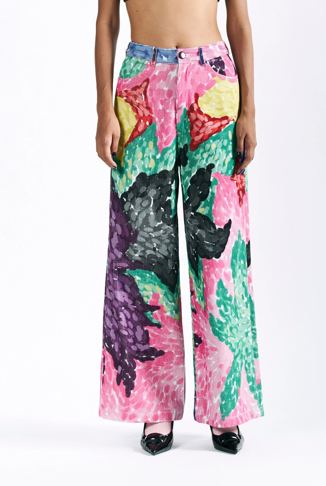 A model is shown from the waist down wearing wide-leg trousers with a vibrant, painterly abstract floral pattern in pink, green, purple, yellow, and black, paired with black pointed shoes against a white studio background.