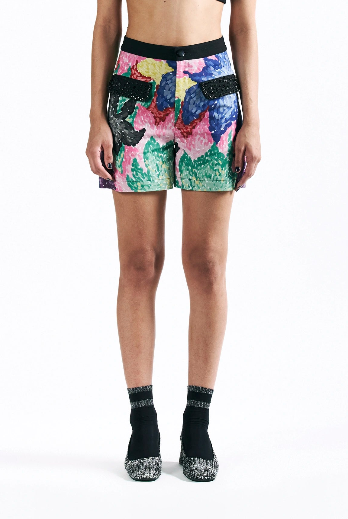 A model is shown from the waist down wearing a pair of colorful abstract-print shorts with black side pockets and black trim, paired with black socks and platform heels, against a plain white background.